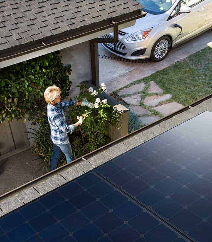 A woman tends to a garden next to a house with solar panels on the roof and an electric car parked in the driveway.