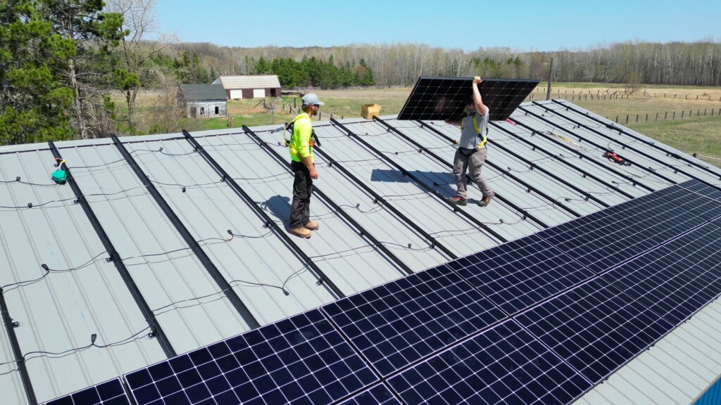 Image of two workers installing solar panels on a metal roof in a rural area with trees and buildings in the background.