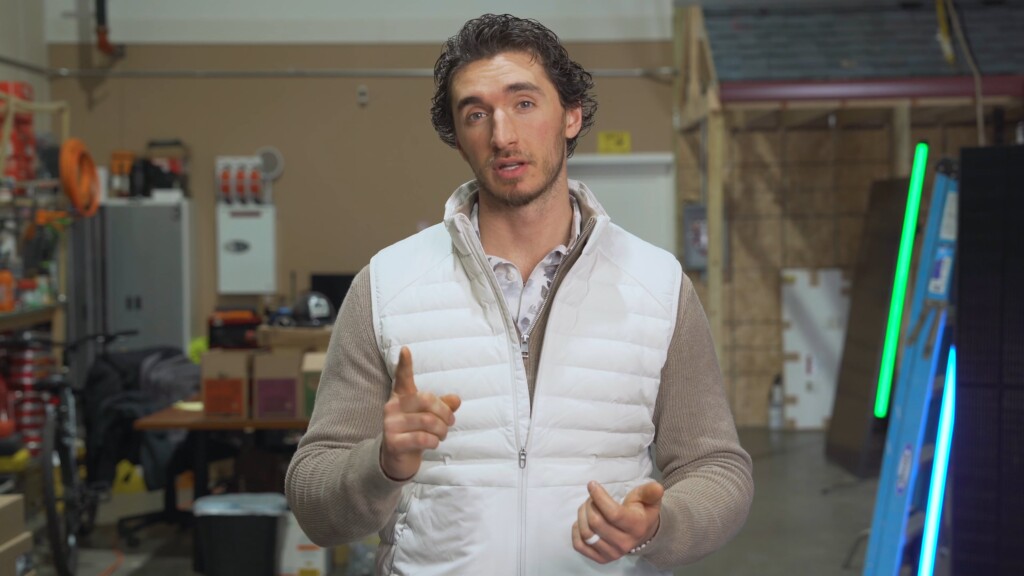 Image of a man wearing a white vest, standing in a workshop or warehouse, speaking to the camera.