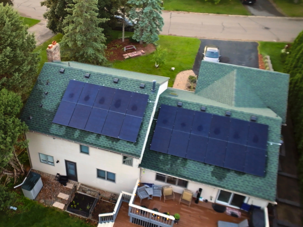 Angled aerial view of a two-story home with green shingle roof and two large solar panel arrays installed, surrounded by trees, deck, and driveway