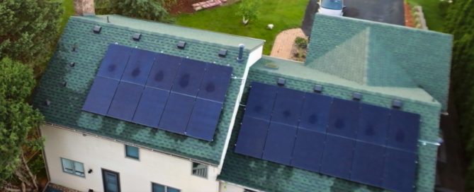 Angled aerial view of a two-story home with green shingle roof and two large solar panel arrays installed, surrounded by trees, deck, and driveway