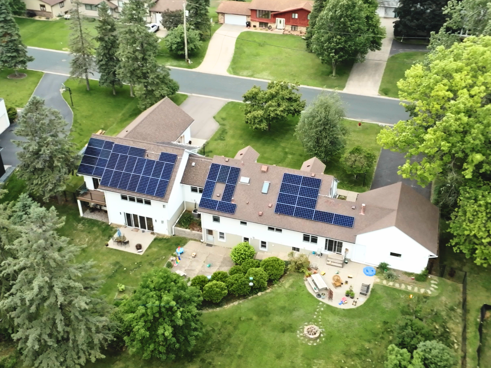 Wide aerial view of a large white two-story house with brown shingle roof and multiple solar panel arrays installed, surrounded by green yard and neighborhood trees Wide aerial view of a large white two-story house with brown shingle roof and multiple solar panel arrays installed, surrounded by green yard and neighborhood trees