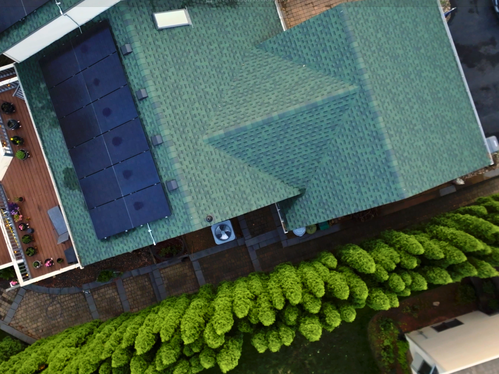 Top-down aerial view of residential roof with green shingles and a solar panel array installed on one side, next to a wooden deck and landscaped yard