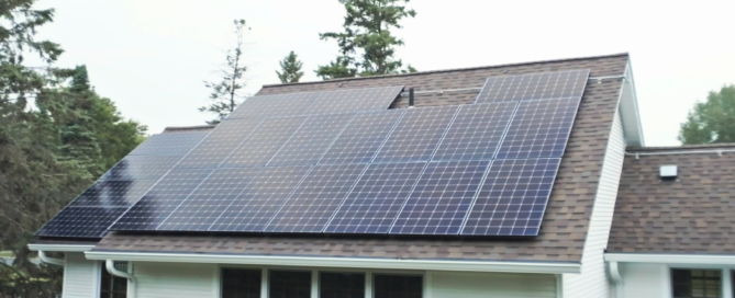 Close-up side view of rooftop solar panel installation on a brown shingle roof of a white house, with trees in the background