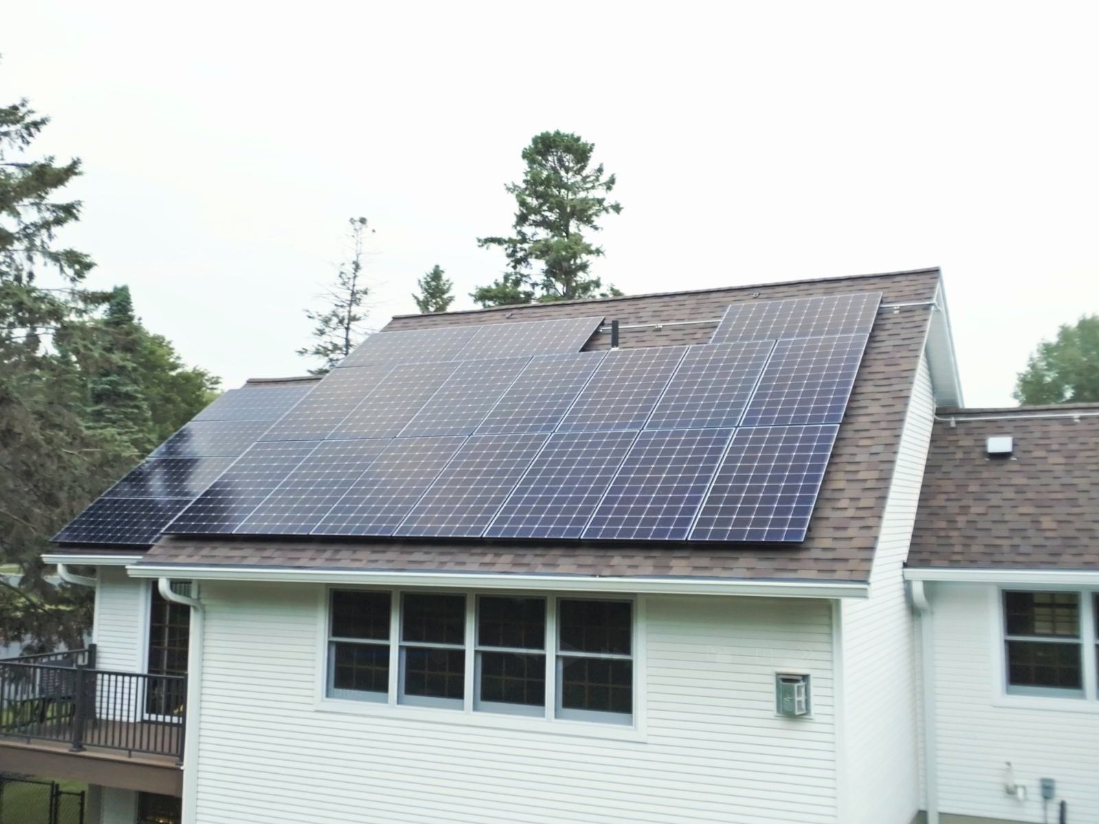 Close-up side view of rooftop solar panel installation on a brown shingle roof of a white house, with trees in the background Close-up side view of rooftop solar panel installation on a brown shingle roof of a white house, with trees in the background