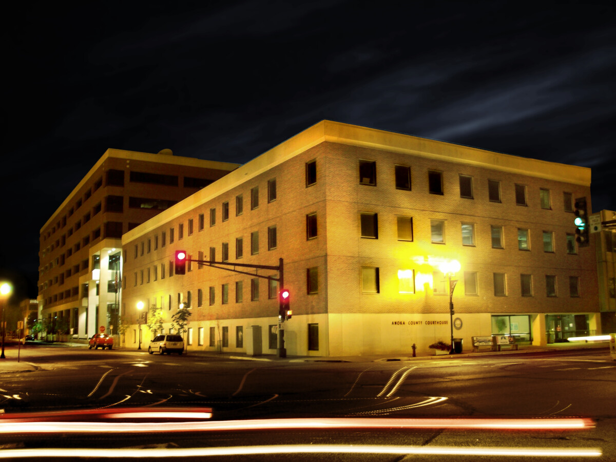Night view of the Anoka County Courthouse in Minnesota, featuring illuminated windows, traffic lights, and light trails from passing cars.