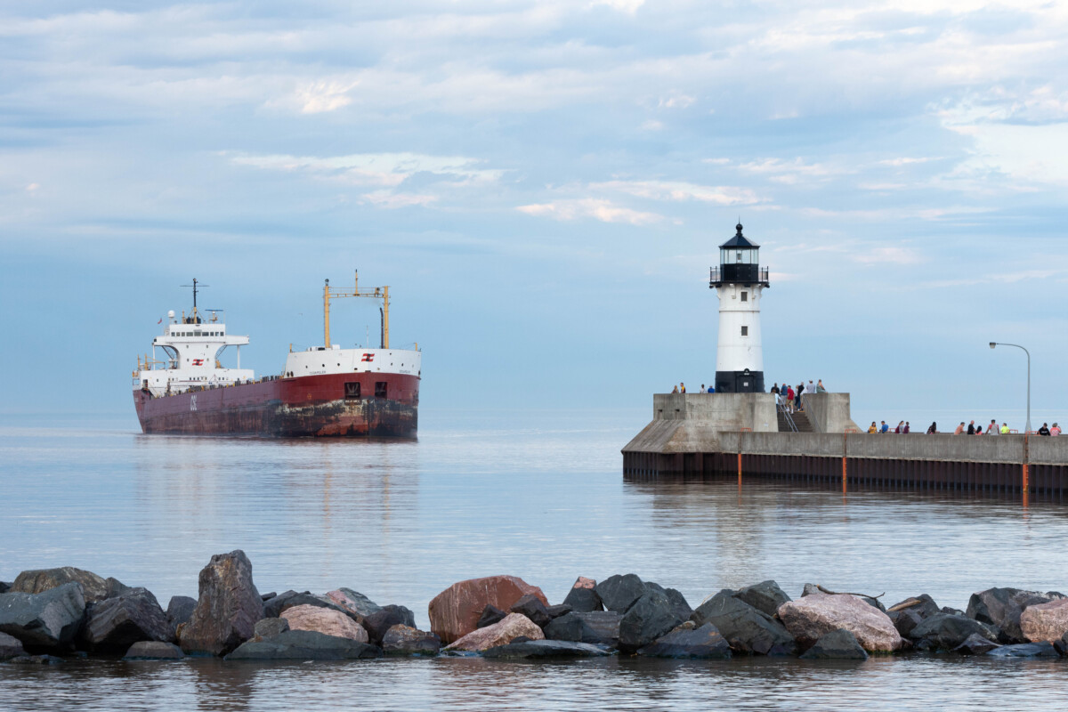 A lighthouse and walkway over water in Duluth, Minnesota with a ship in the background during the day. A lighthouse and walkway over water in Duluth, Minnesota with a ship in the background during the day.