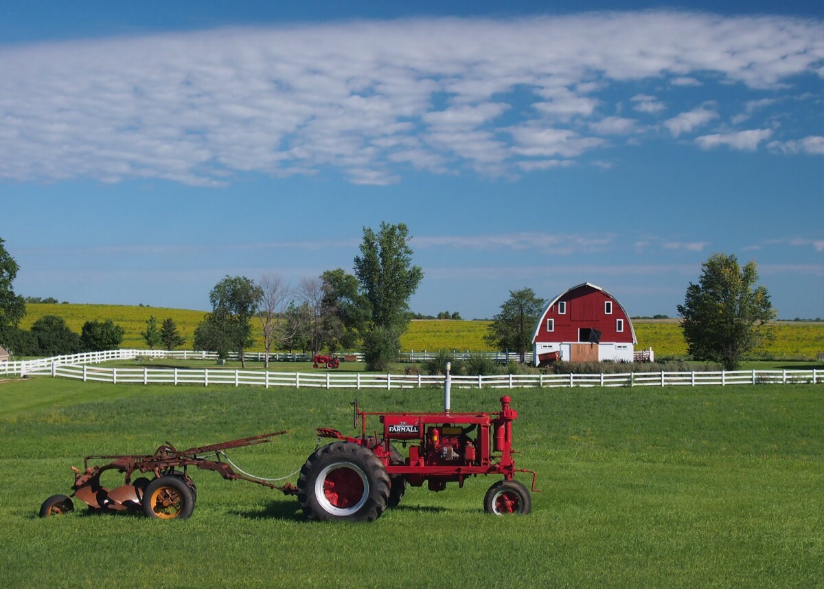 Red barn and tractor surrounded by green grass and trees in front of a cloudy blue sky.