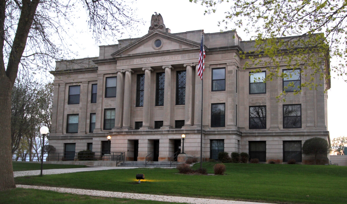 A front shot of the Lincoln County Courthouse in Minnesota with trees and an American flag in the forefront during the day.