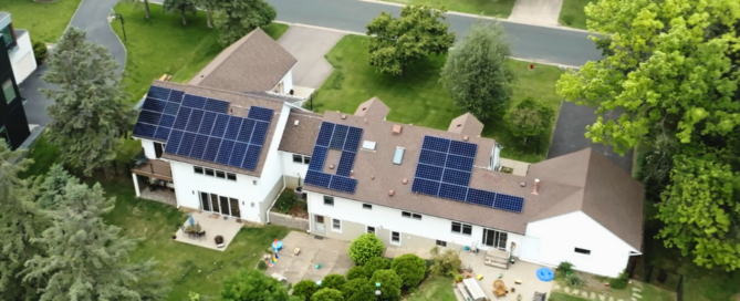 Wide aerial view of a large white two-story house with brown shingle roof and multiple solar panel arrays installed, surrounded by green yard and neighborhood trees