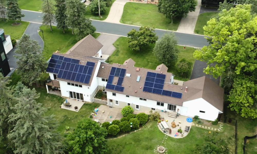 Wide aerial view of a large white two-story house with brown shingle roof and multiple solar panel arrays installed, surrounded by green yard and neighborhood trees
