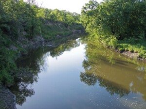 Calm river winding through verdant banks of shrubs and trees, reflecting green foliage on a sunny day Calm river winding through verdant banks of shrubs and trees, reflecting green foliage on a sunny day