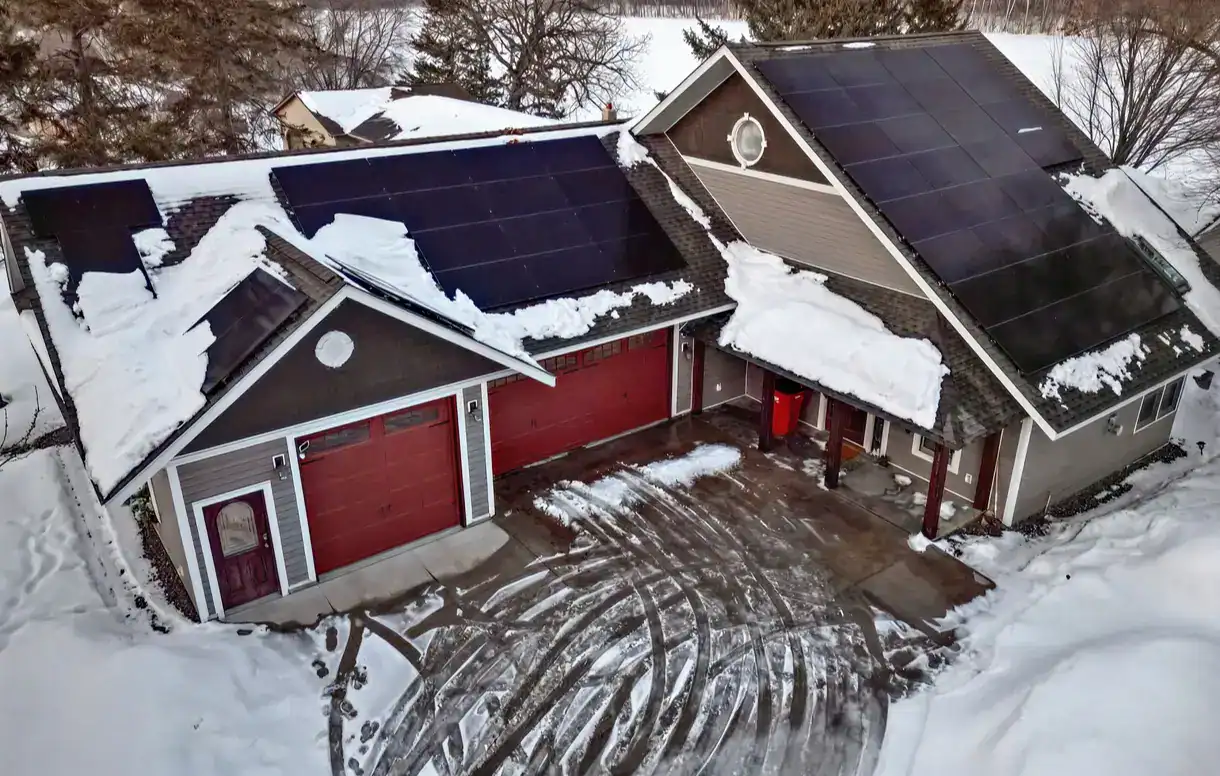 A snowy aerial view of a residential home with solar panels installed on the roofs of both the main house and attached garage.