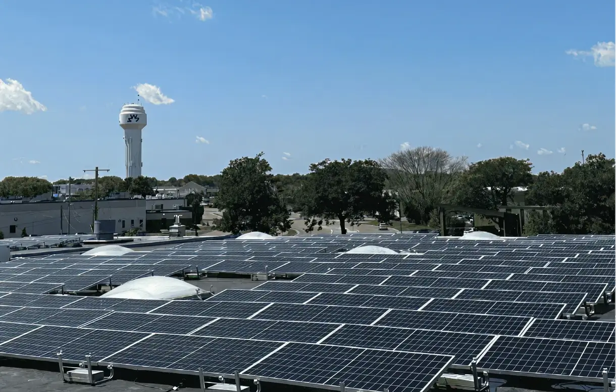 Rooftop covered with rows of solar panels under a clear blue sky, with a water tower and trees in the background.