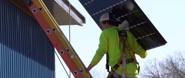Wolf River Electric technician installing rooftop solar panels on a residential home using a ladder and safety gear during a solar energy system installation.