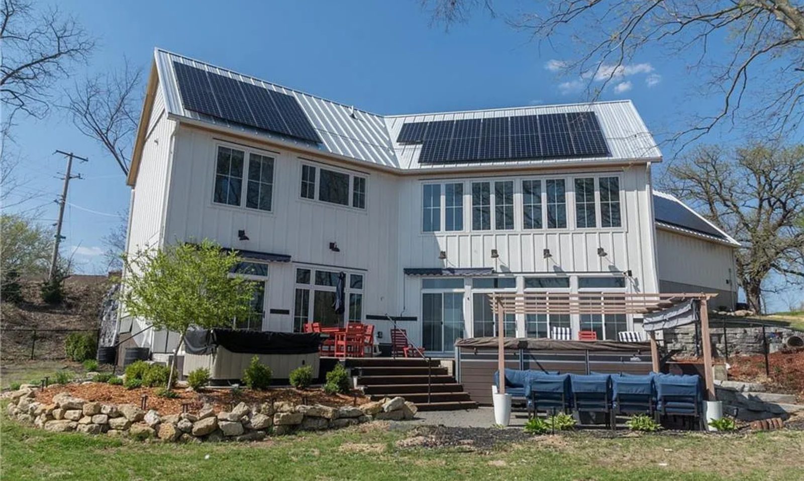 Modern farmhouse in Wisconsin with rooftop solar panels installed on a white metal roof, surrounded by landscaped yard, patio seating, and outdoor living space.
