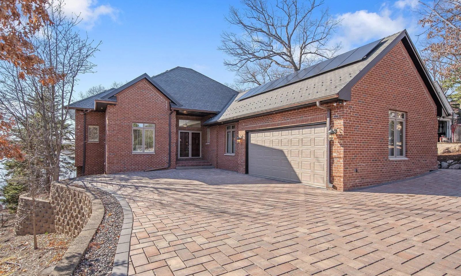 Brick home in Wisconsin featuring rooftop solar panels on an asphalt shingle roof, with paved driveway, garage, and mature trees in the background.